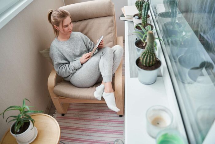 Blonde girl reading book while sitting on beige armchair in apartment on balcony