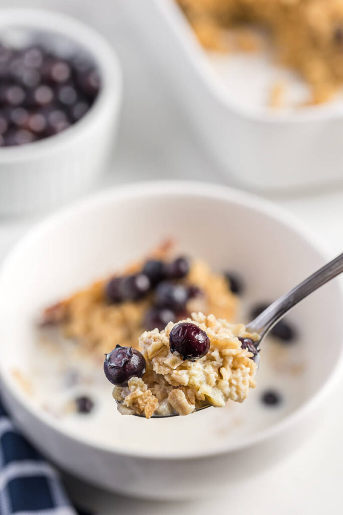 Blueberry Baked Oatmeal on a spoon. White bowl with Baked oatmeal and fresh blueberries